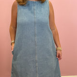A woman wearing a sleeveless light blue corduroy dress with side pockets, standing against a solid pink wall.