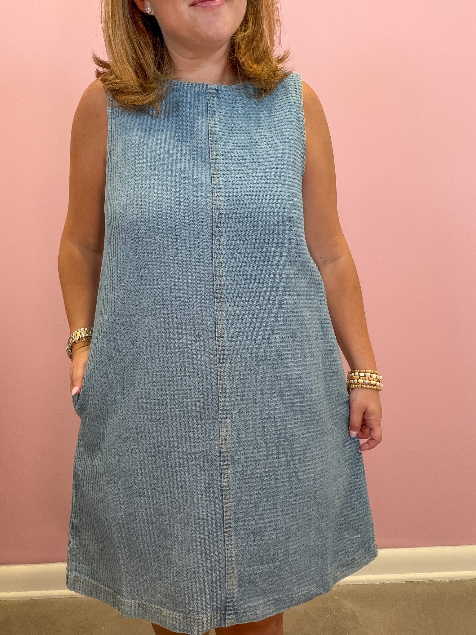 A woman wearing a sleeveless light blue corduroy dress with side pockets, standing against a solid pink wall.