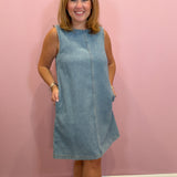 A woman modeling a sleeveless light blue corduroy dress with side pockets, standing against a solid pink wall.