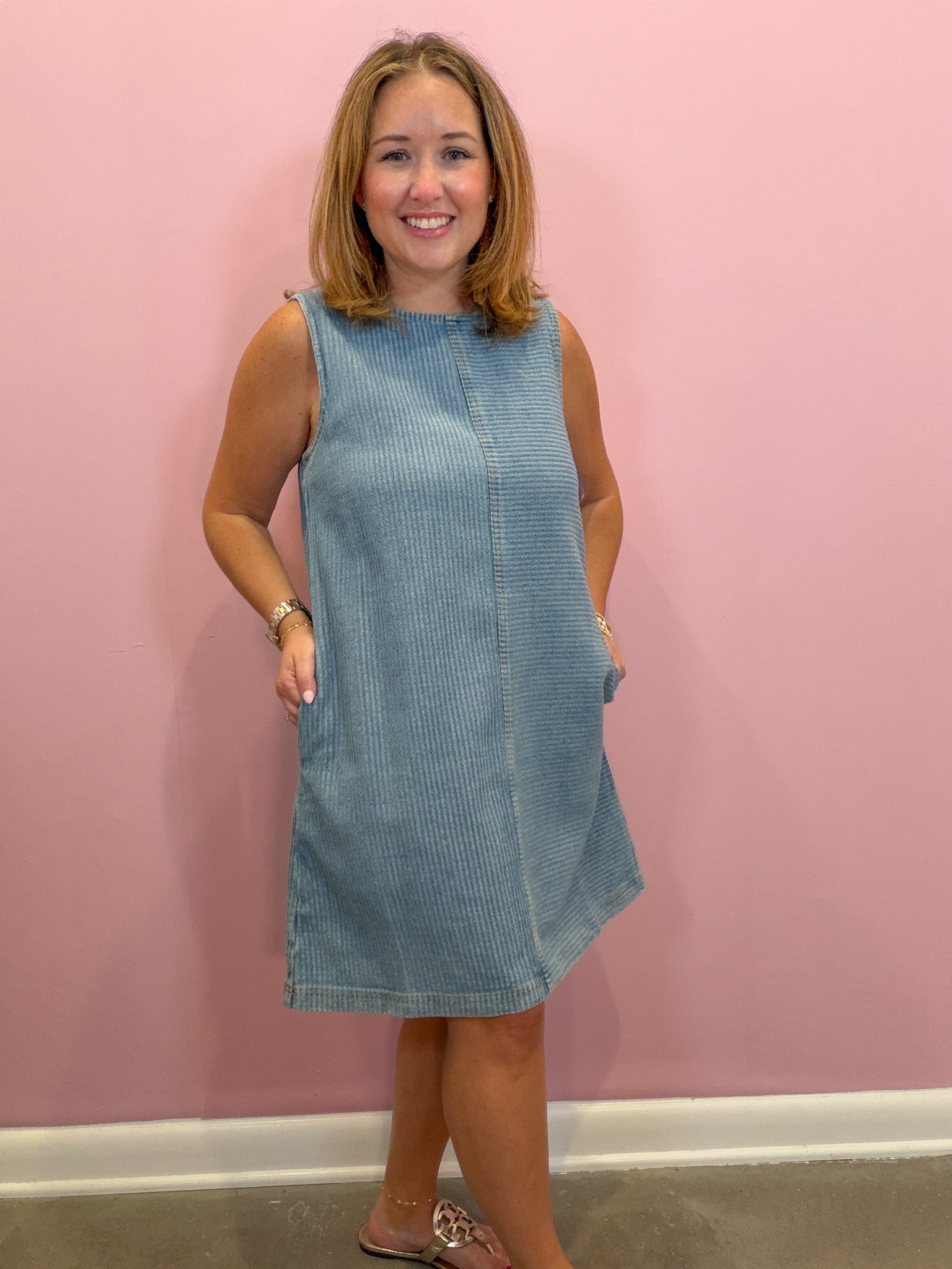 A woman modeling a sleeveless light blue corduroy dress with side pockets, standing against a solid pink wall.