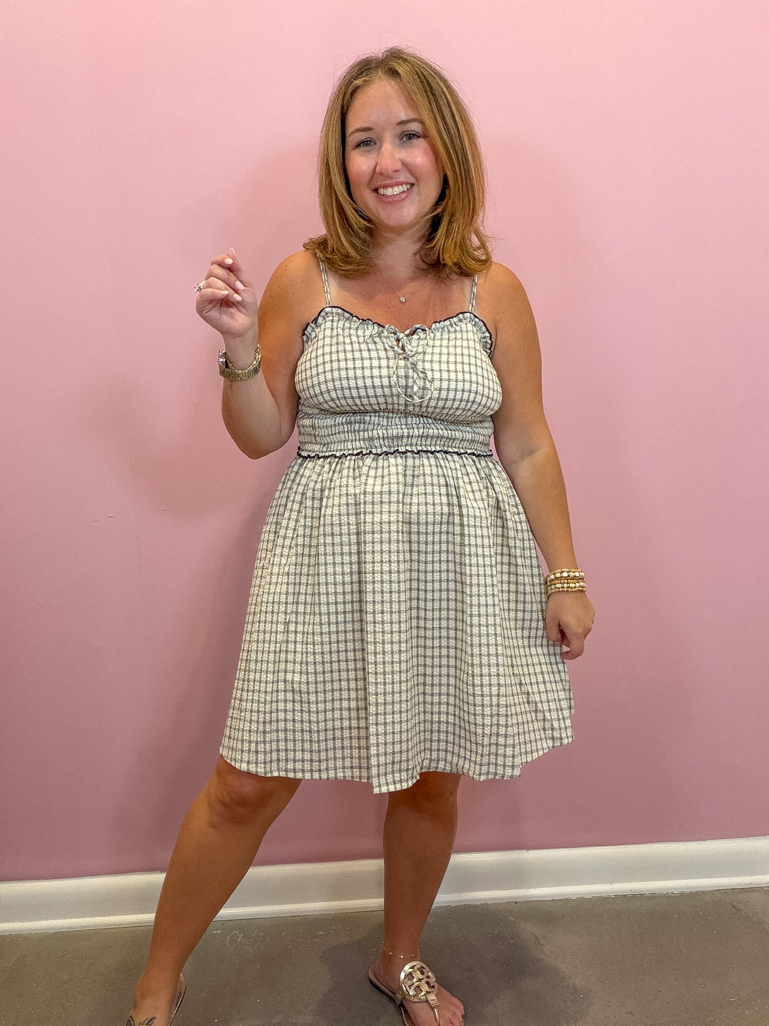 A woman modeling a sleeveless A-line dress with a green and white gingham pattern, standing against a solid pink wall.