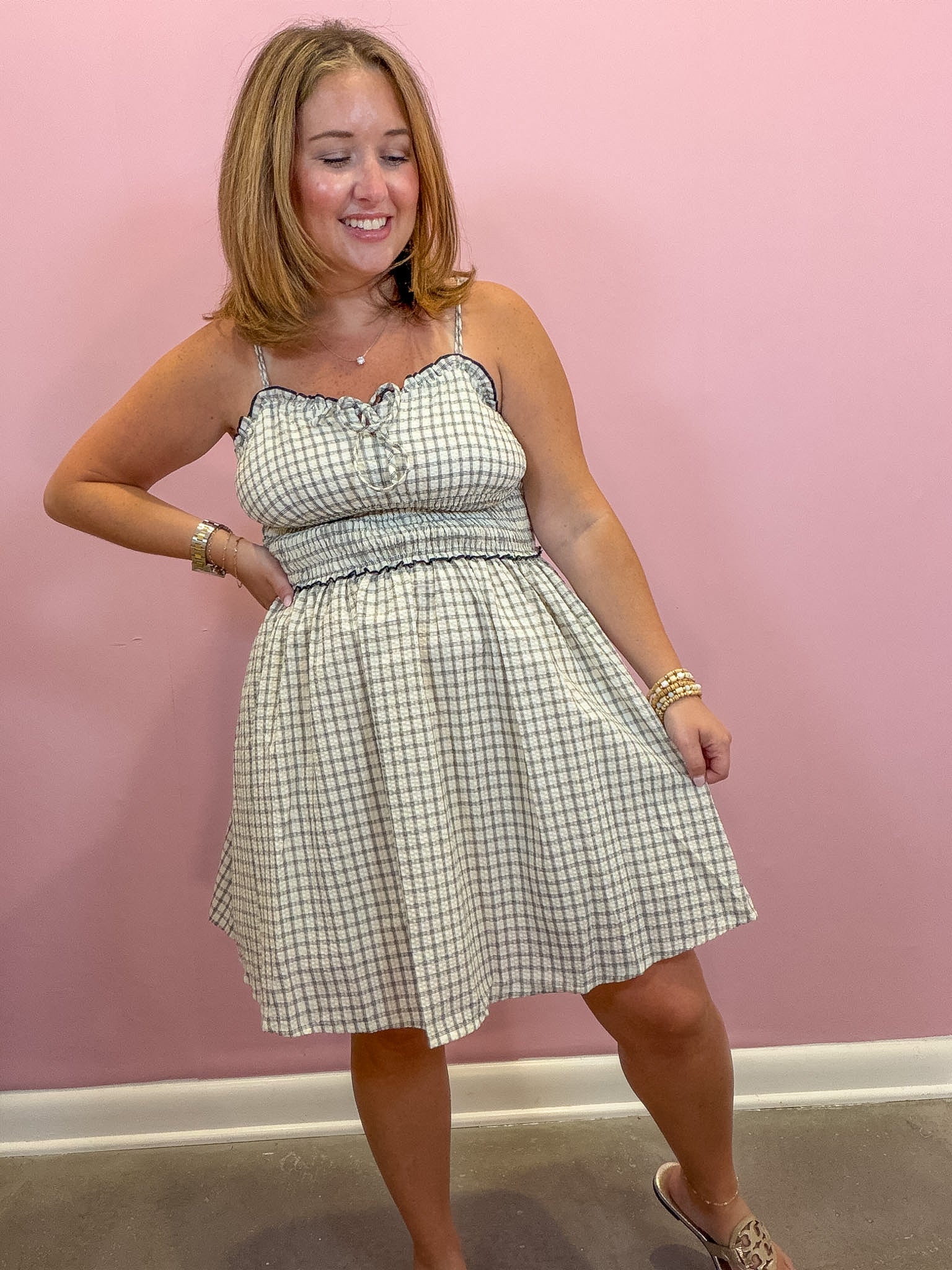 A woman modeling a sleeveless A-line dress with a green and white gingham pattern, featuring a smocked bodice and spaghetti straps, posing against a solid pink wall.