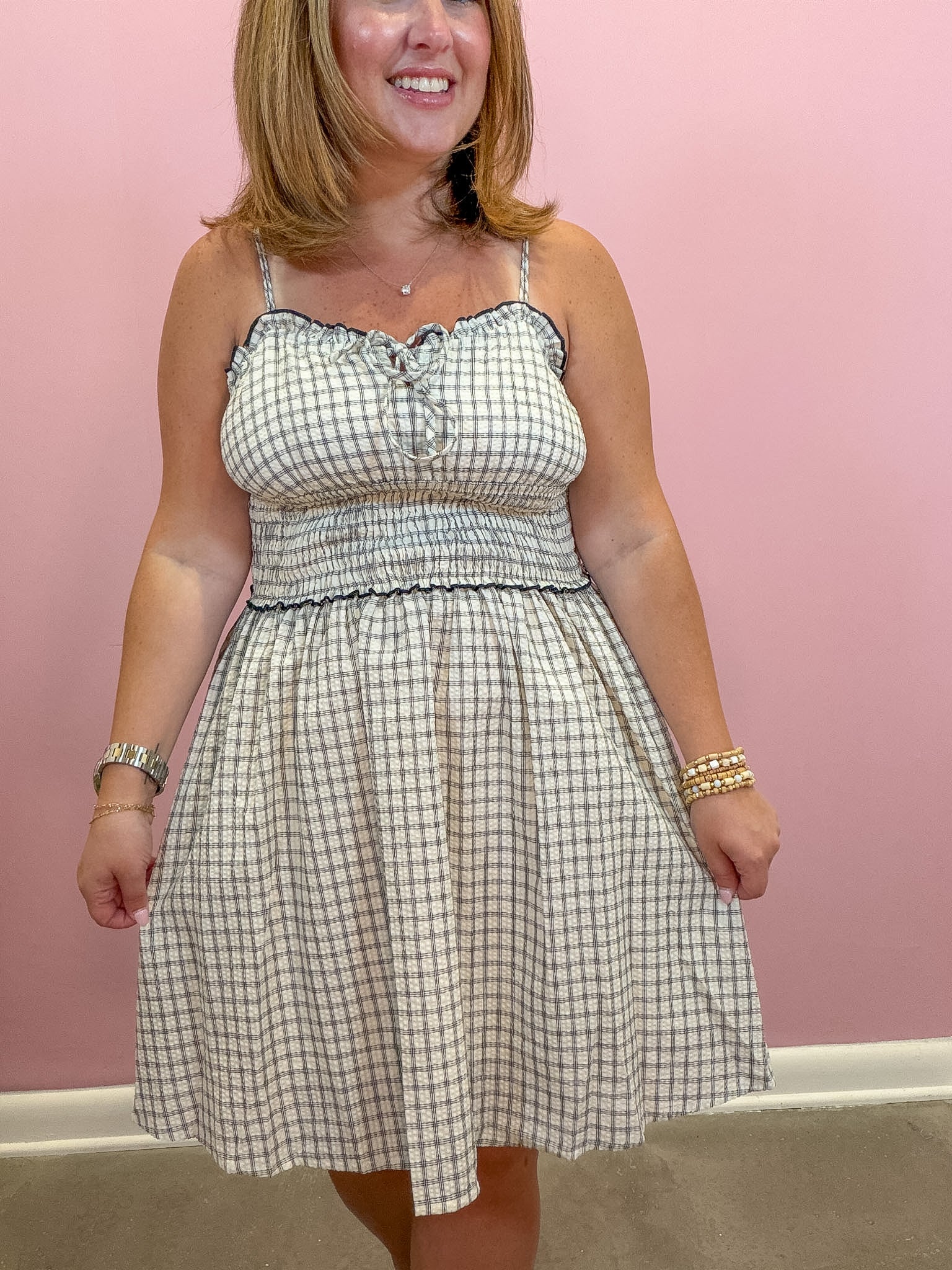 A woman wearing a sleeveless, smocked A-line dress with a beige and navy blue gingham pattern, standing against a solid pink wall.