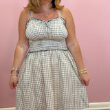 A woman wearing a sleeveless, smocked A-line dress with a beige and navy blue gingham pattern, standing against a solid pink wall.