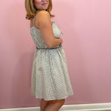 A woman modeling a sleeveless, knee-length A-line dress with a white and navy blue gingham pattern, standing against a solid pink wall.