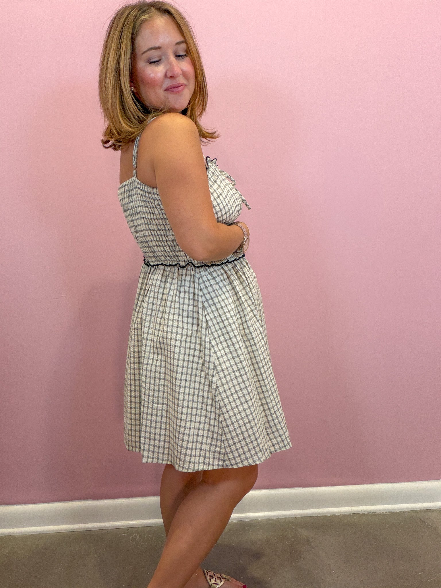 A woman modeling a sleeveless, knee-length A-line dress with a white and navy blue gingham pattern, standing against a solid pink wall.