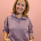 A smiling woman with shoulder-length brown hair wearing a dusty purple quarter-zip sweatshirt and matching sweatpants, standing against a solid pink background.