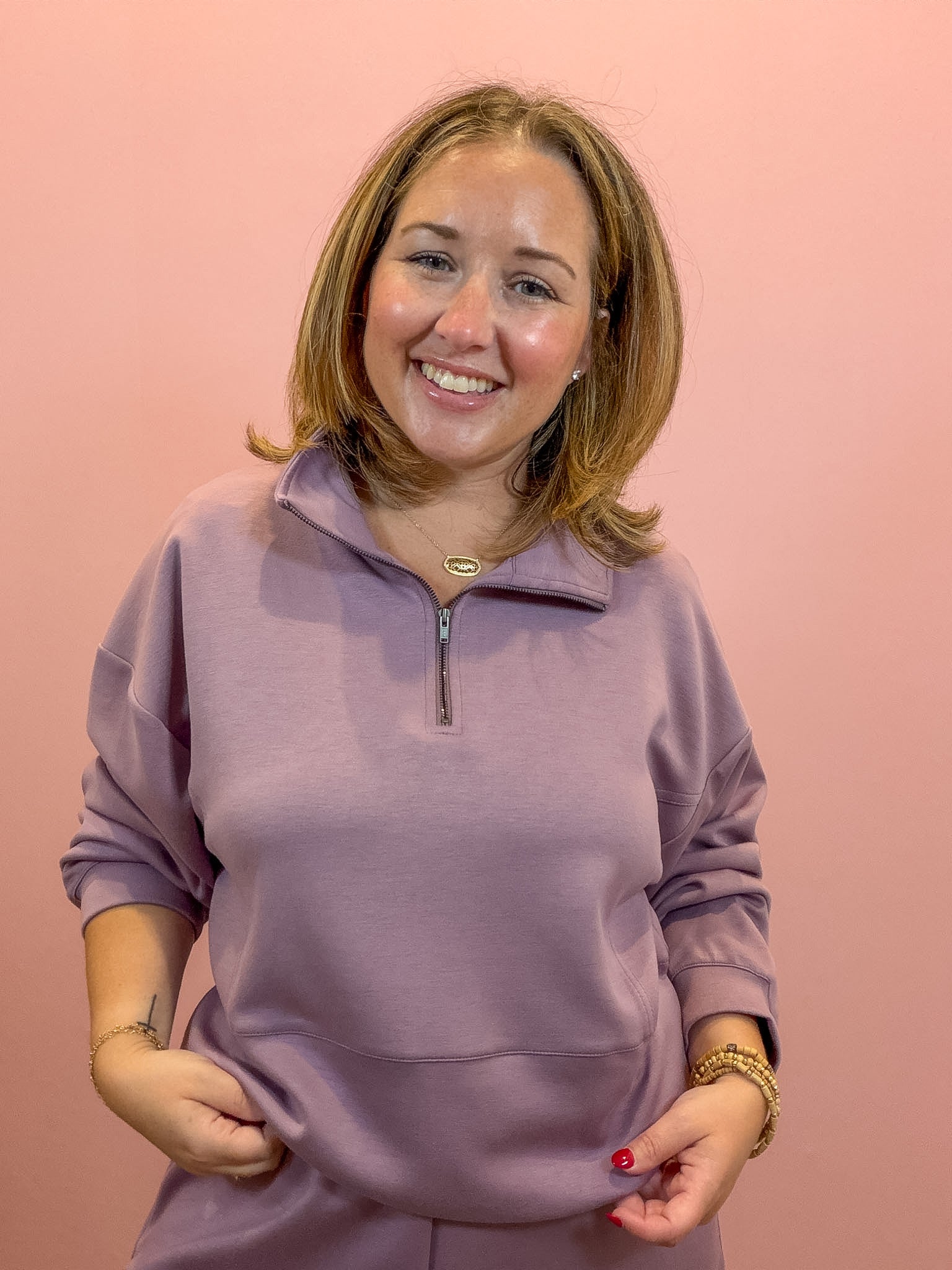 A smiling woman with shoulder-length brown hair wearing a dusty purple quarter-zip sweatshirt and matching sweatpants, standing against a solid pink background.