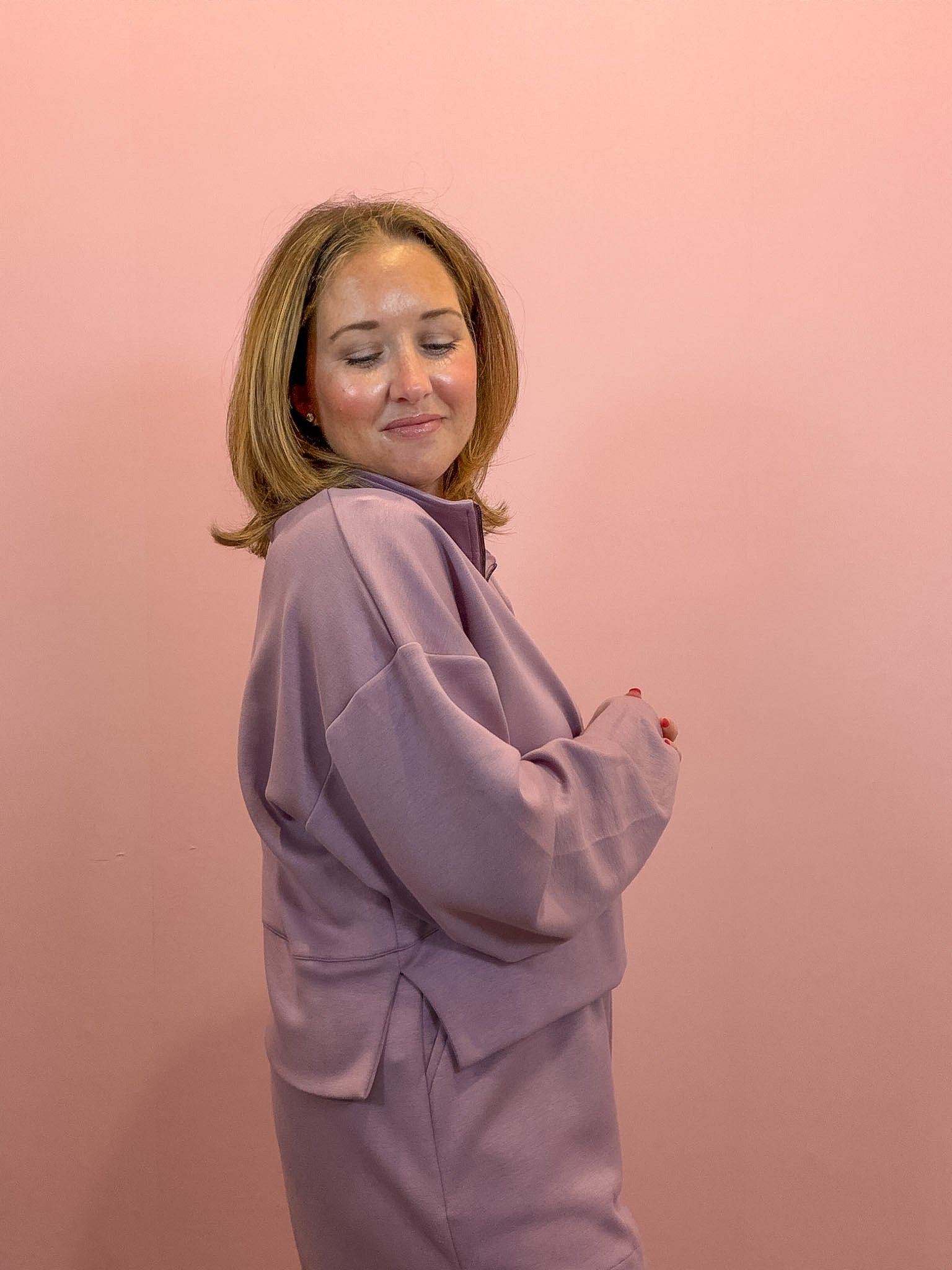 A woman with shoulder-length blonde hair wearing a lavender purple fleece jacket and matching pants, posing against a solid pink background.