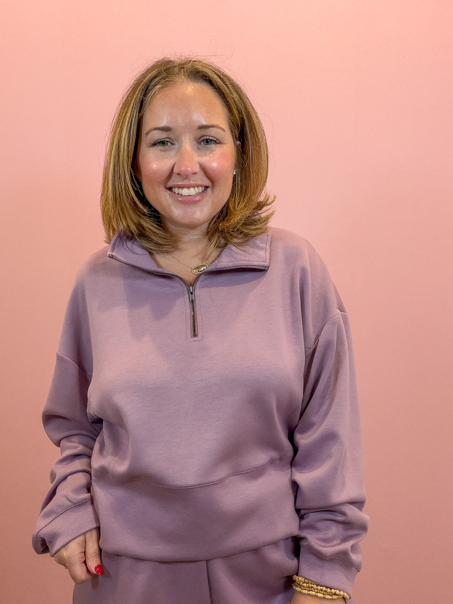 A smiling woman with shoulder-length blonde hair wearing a dusty purple quarter-zip sweatshirt and matching sweatpants, standing against a solid pink background.
