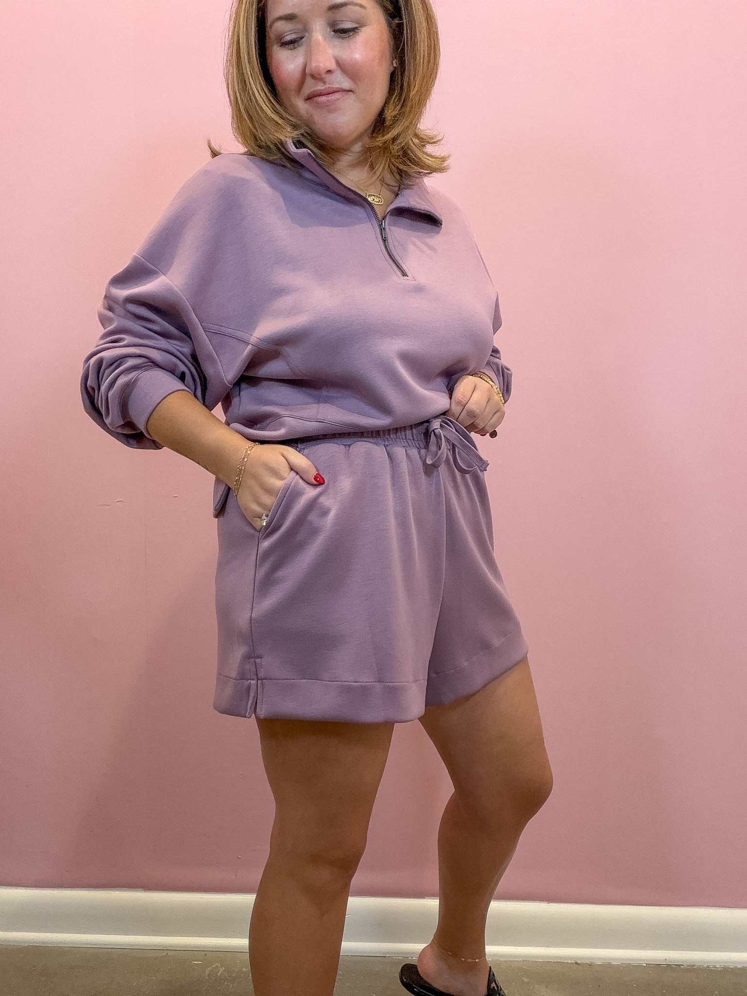 A woman modeling a matching lavender lounge set consisting of a half-zip sweatshirt and coordinating shorts, standing against a solid pink wall.