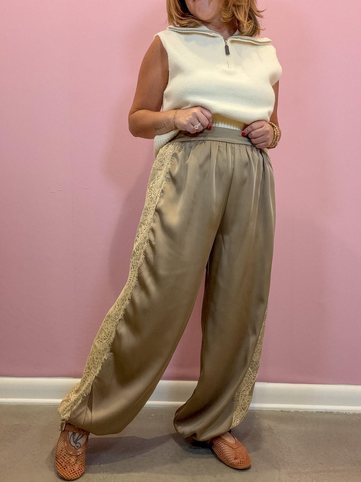 A person wearing wide-leg beige satin pants with lace side panels, a cream sleeveless quarter-zip top, and brown woven shoes, standing against a pink wall.