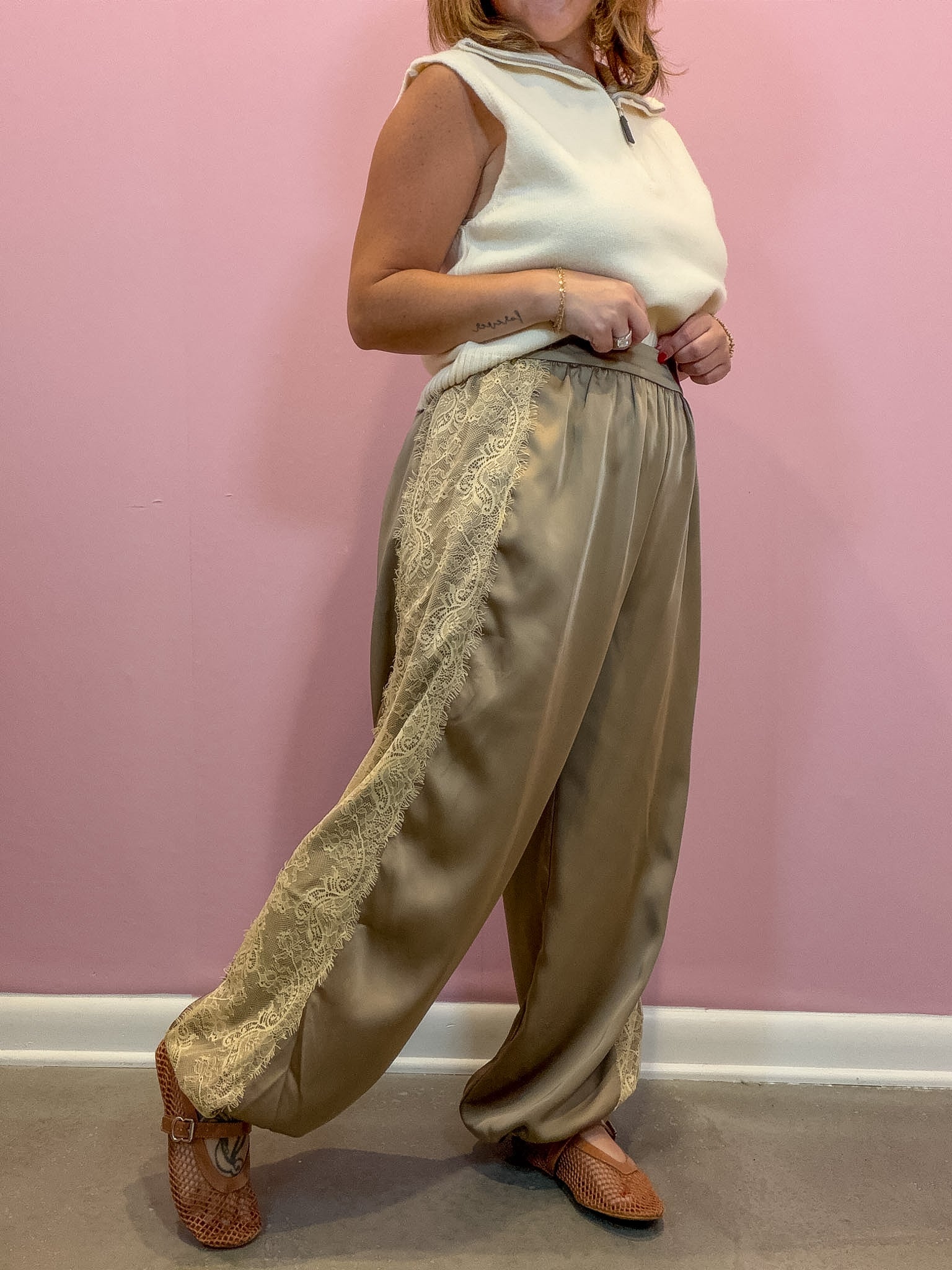 A person wearing wide-leg satin pants with lace trim, a cream sleeveless top, and brown woven shoes, standing against a pink wall.