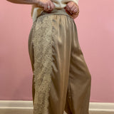 A person wearing wide-leg taupe satin pants with a vertical cream lace panel down the left leg, paired with a cream knit top, standing against a pink wall.