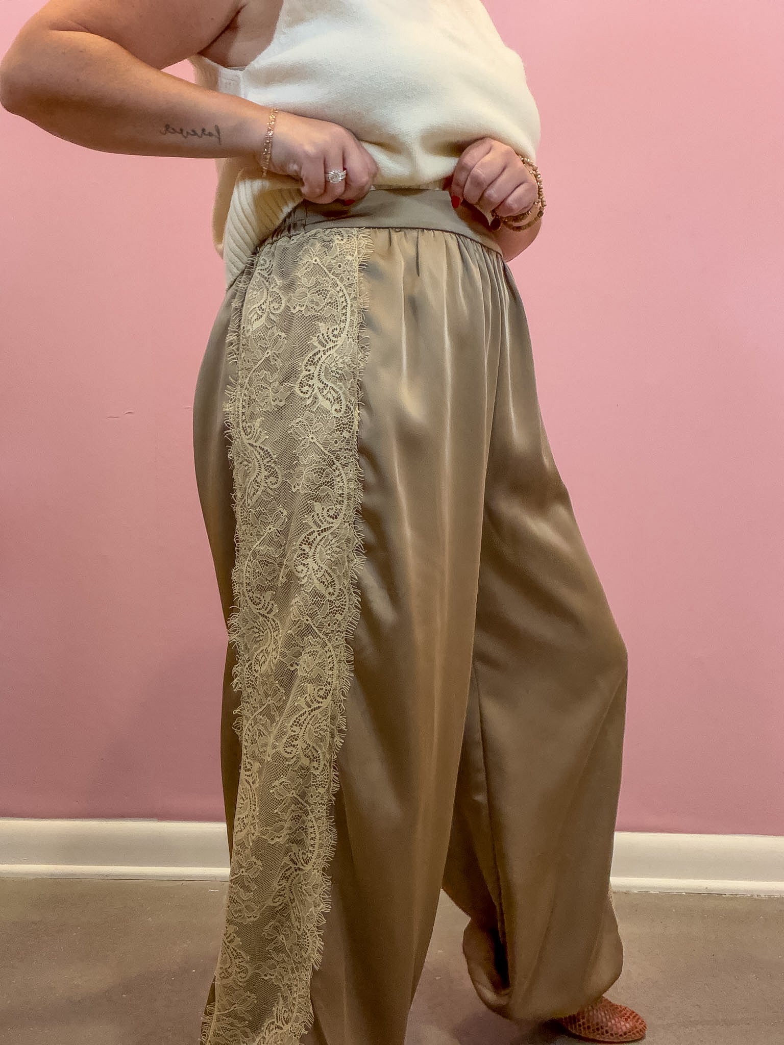 A person wearing wide-leg taupe satin pants with a vertical cream lace panel down the left leg, paired with a cream knit top, standing against a pink wall.