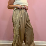 A person wearing loose, wide-leg taupe satin pants with a beige lace panel on the side, paired with a cream sleeveless top and brown shoes, standing against a pink wall.