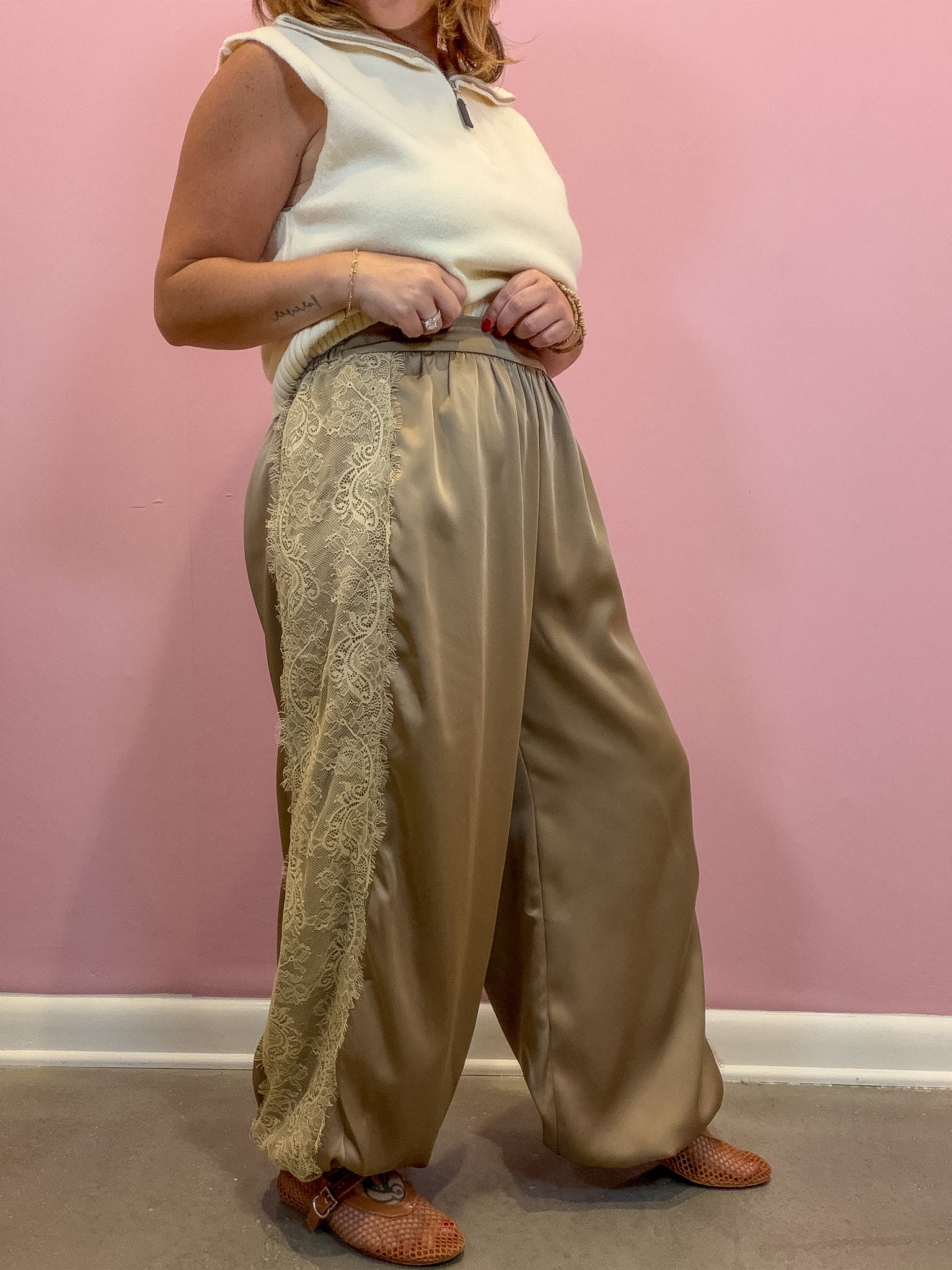 A person wearing loose, wide-leg taupe satin pants with a beige lace panel on the side, paired with a cream sleeveless top and brown shoes, standing against a pink wall.
