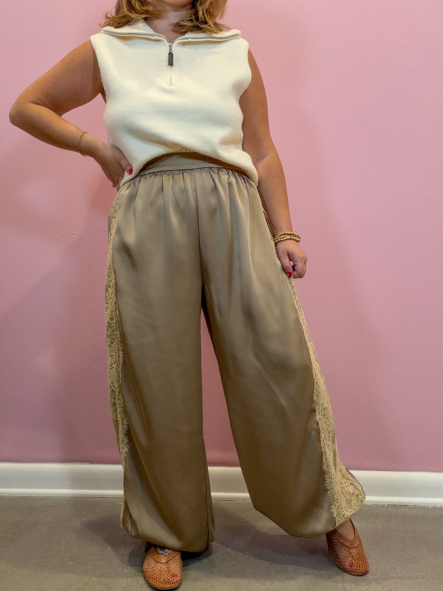 A model wearing wide-leg taupe satin pants with beige lace side panels and a cream sleeveless quarter-zip top, standing against a pink wall.