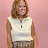 A woman with shoulder-length blonde hair smiling in front of a solid pink background. She is wearing a cream-colored sleeveless quarter-zip knit top and brown pleated trousers.