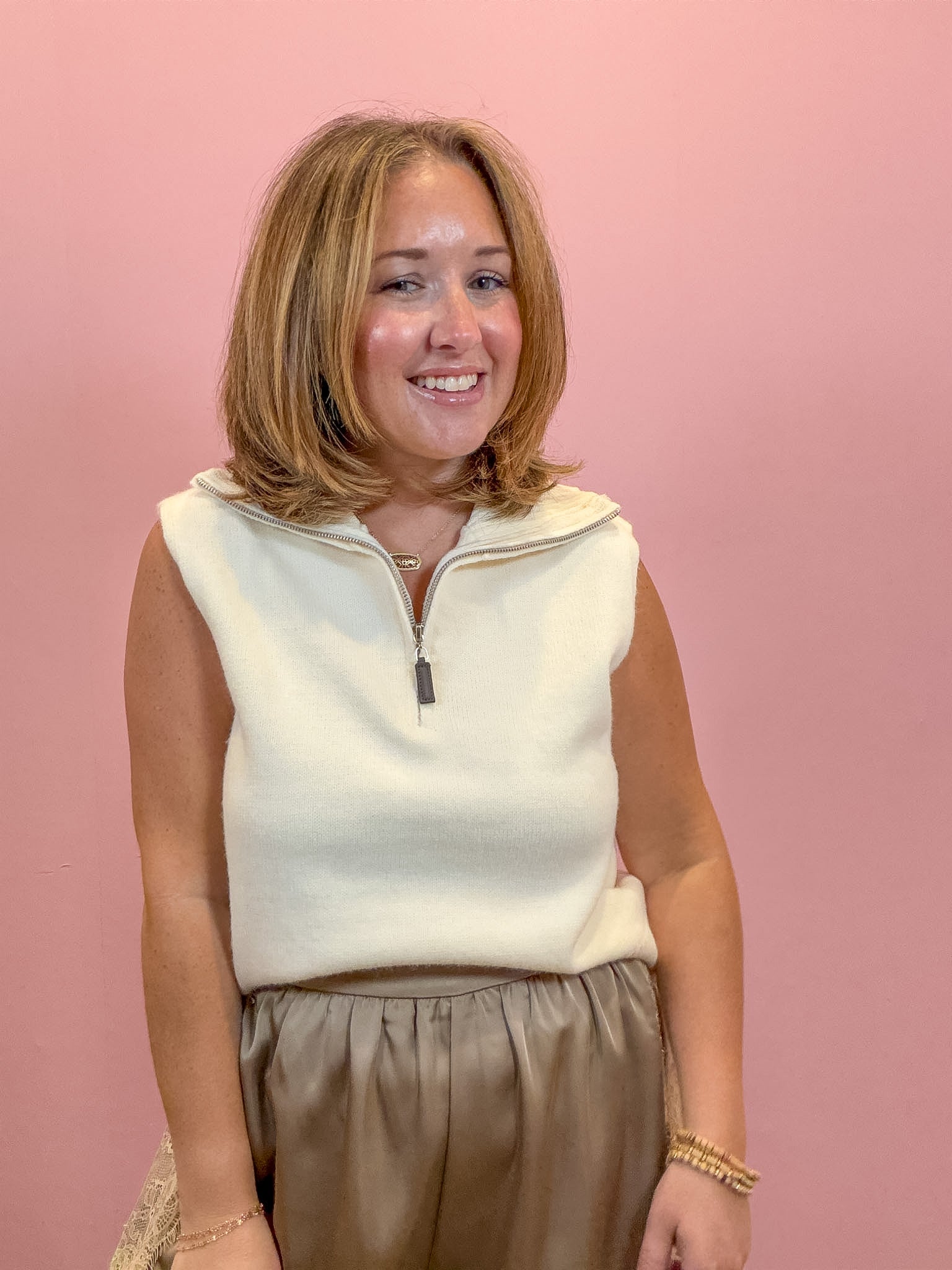 A woman with shoulder-length blonde hair smiling in front of a solid pink background. She is wearing a cream-colored sleeveless quarter-zip knit top and brown pleated trousers.