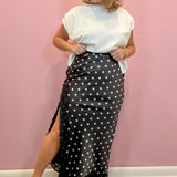 A woman modeling a black wrap-style midi skirt with white polka dots and a side slit, paired with a white top and black thong sandals, standing against a pink wall.
