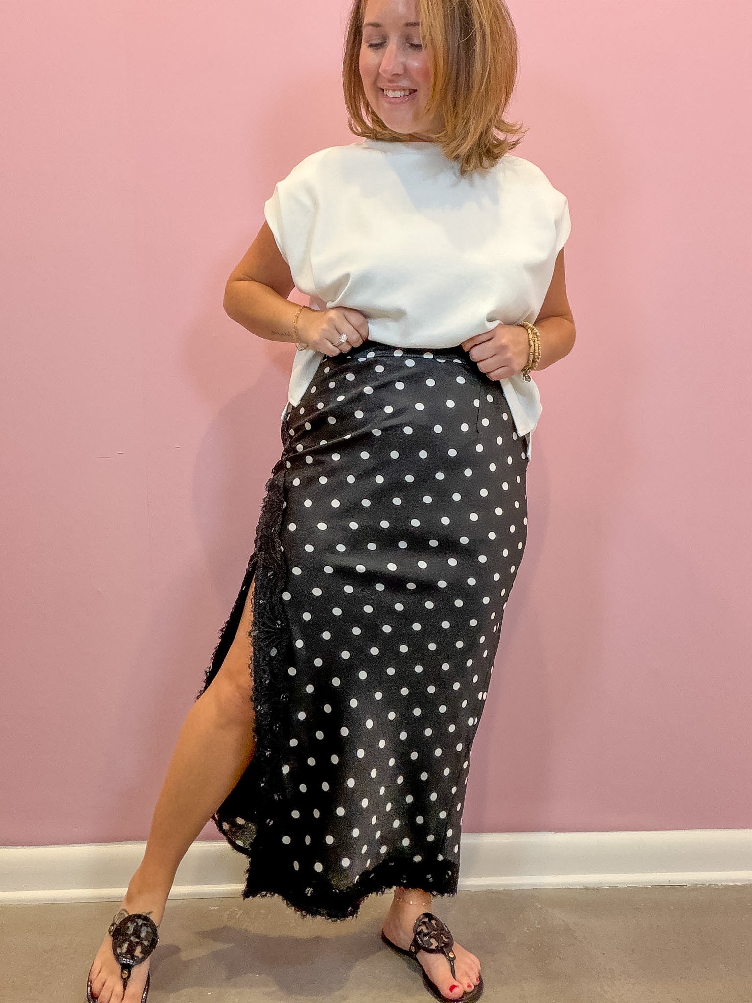 A woman modeling a black wrap-style midi skirt with white polka dots and a side slit, paired with a white top and black thong sandals, standing against a pink wall.