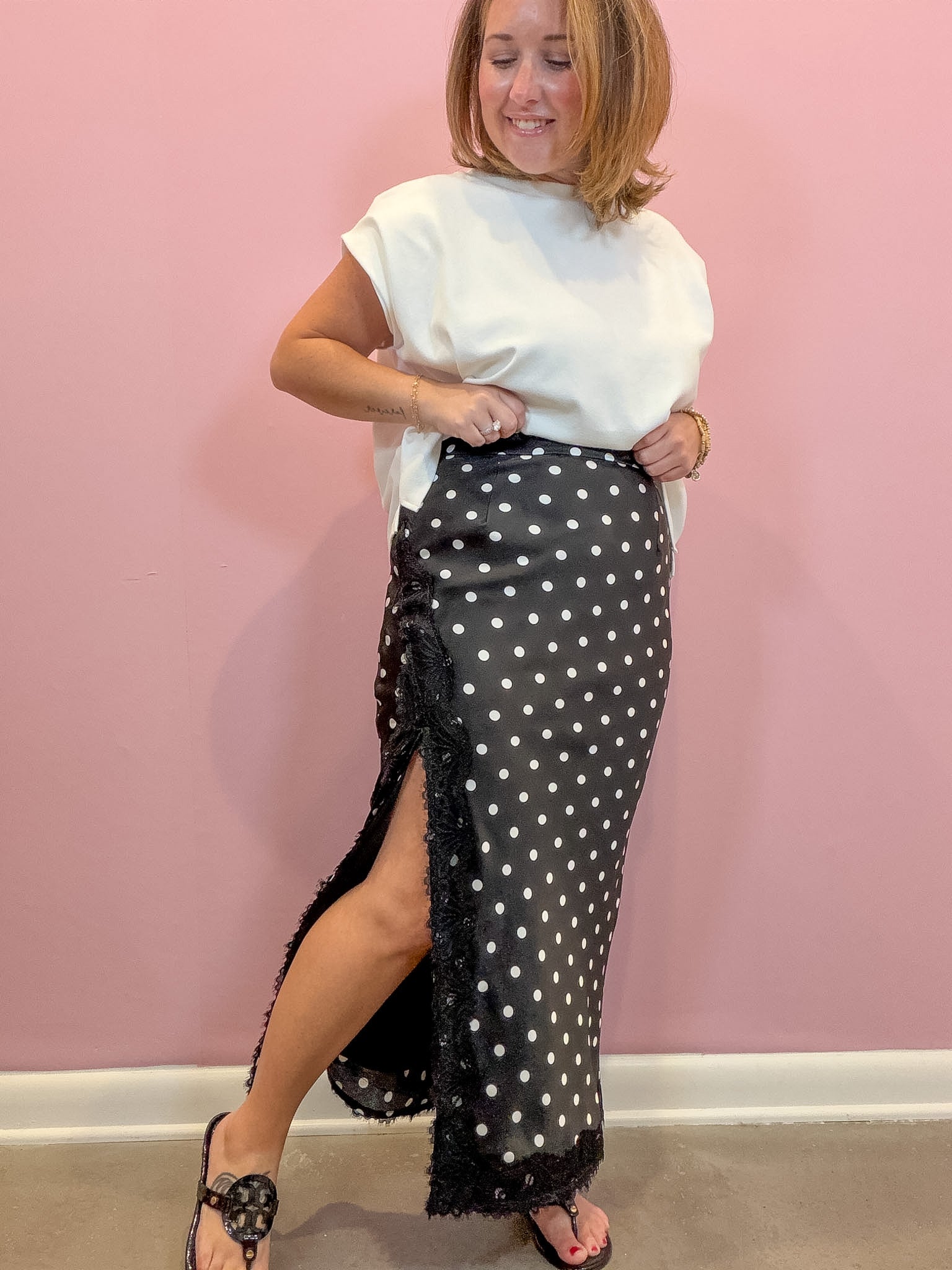 A woman modeling a black midi skirt with white polka dots and a fringed hem, paired with a white t-shirt and black sandals, standing against a pink wall.