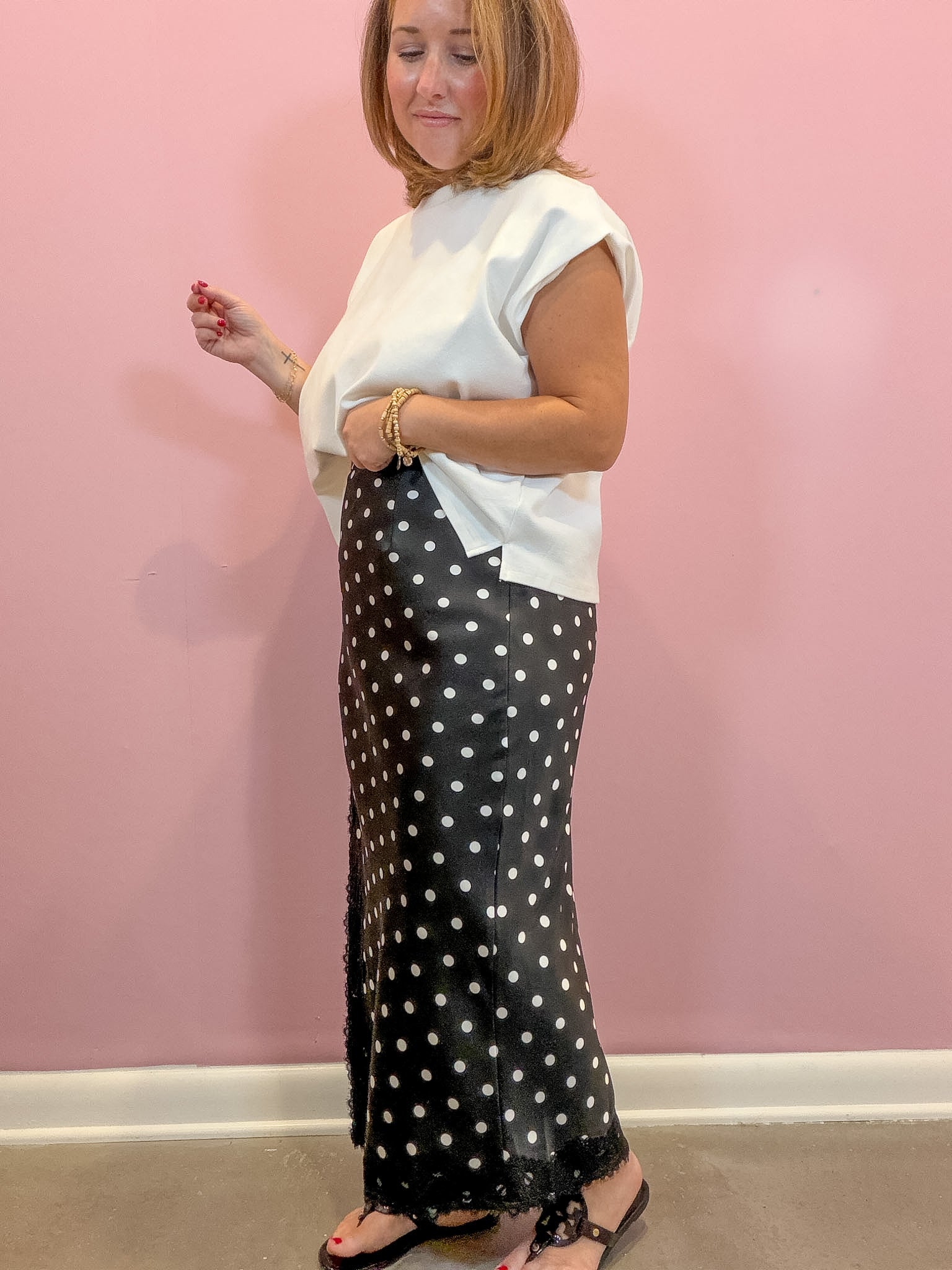 A woman modeling a white sleeveless top and a black maxi skirt with white polka dots, standing against a pink wall.