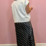 A woman shown from the back wearing a white sleeveless top and a black maxi skirt with white polka dots, standing against a pink wall.