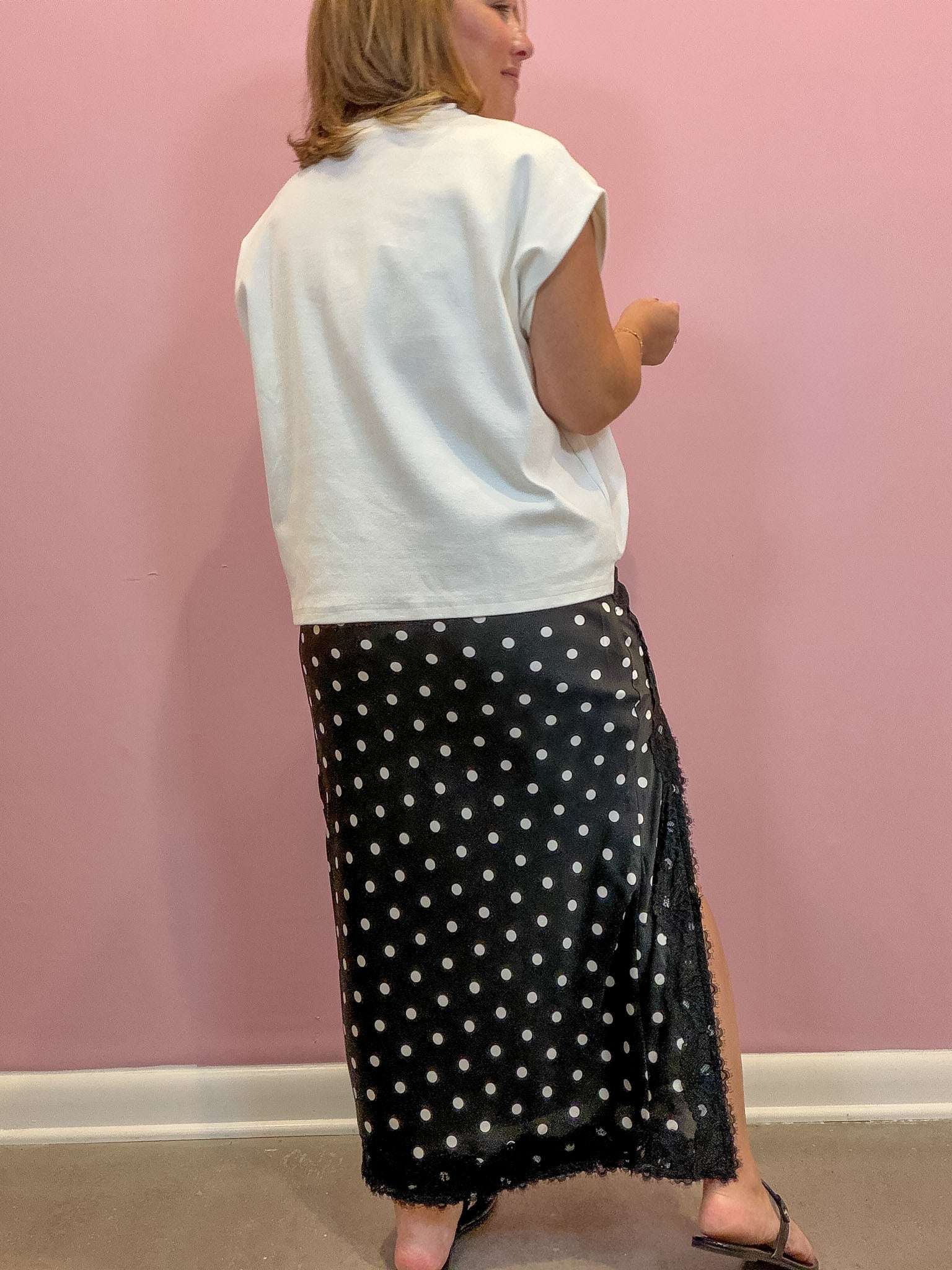 A woman shown from the back wearing a white sleeveless top and a black maxi skirt with white polka dots, standing against a pink wall.