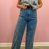 A person wearing a light gray sleeveless button-front cardigan and high-waisted wide-leg blue denim jeans, standing against a pink wall.