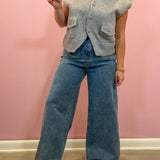 A woman wearing a light gray sleeveless button-front knit vest and wide-leg blue jeans, standing against a pink wall.
