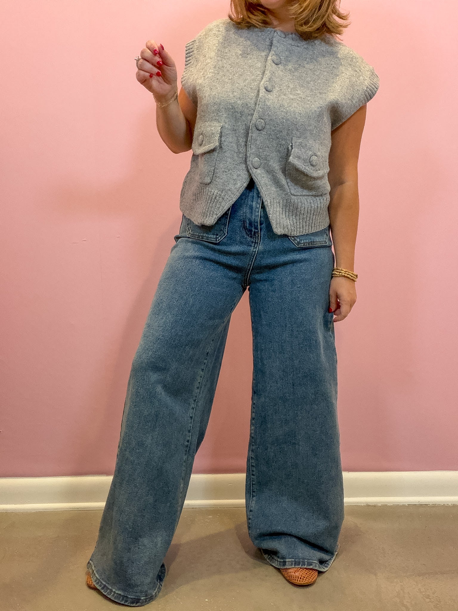 A woman wearing a light gray sleeveless button-front knit vest and wide-leg blue jeans, standing against a pink wall.
