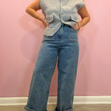 A woman modeling a light gray sleeveless button-front knit vest with front pockets, paired with wide-leg blue jeans and brown shoes, standing against a pink wall.