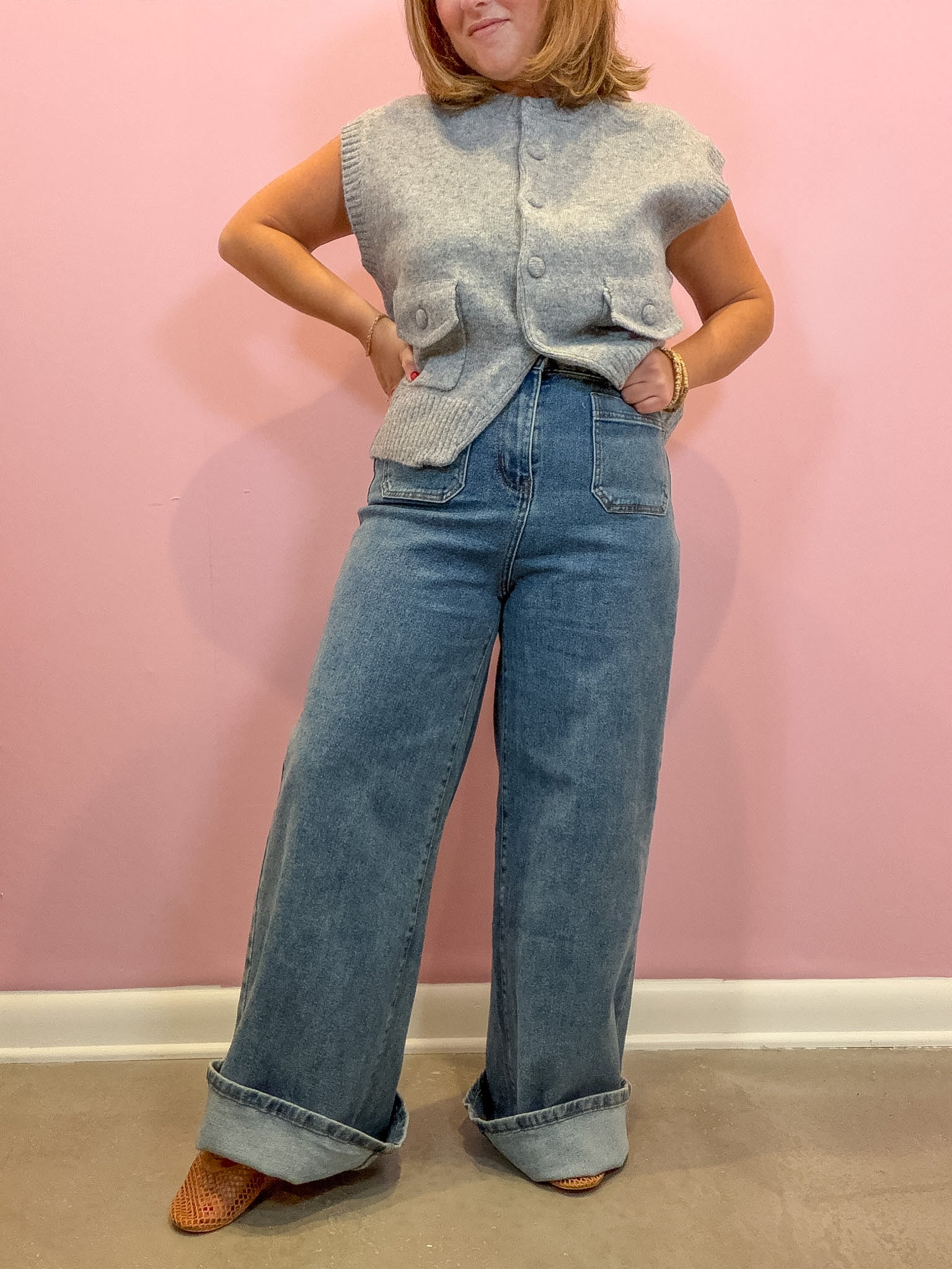 A woman modeling a light gray sleeveless button-front knit vest with front pockets, paired with wide-leg blue jeans and brown shoes, standing against a pink wall.