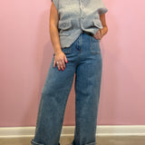A woman modeling a light gray sleeveless button-front cardigan vest over a light blue top, paired with wide-leg light blue denim jeans and brown shoes, standing against a pink wall.