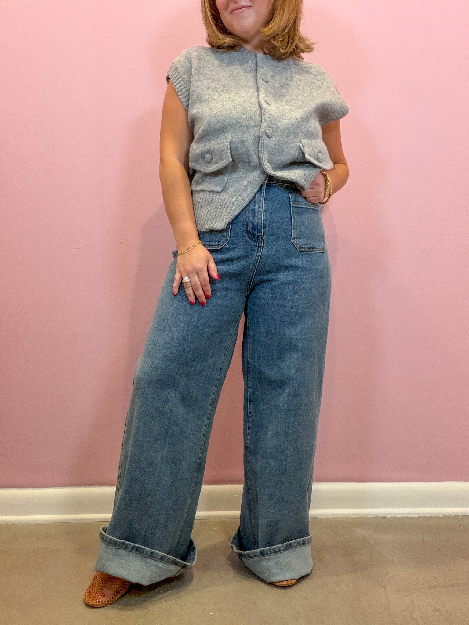 A woman modeling a light gray sleeveless button-front cardigan vest over a light blue top, paired with wide-leg light blue denim jeans and brown shoes, standing against a pink wall.
