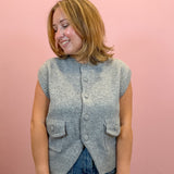 A woman wearing a gray sleeveless button-front knit vest over a light blue top, standing against a solid pink background.