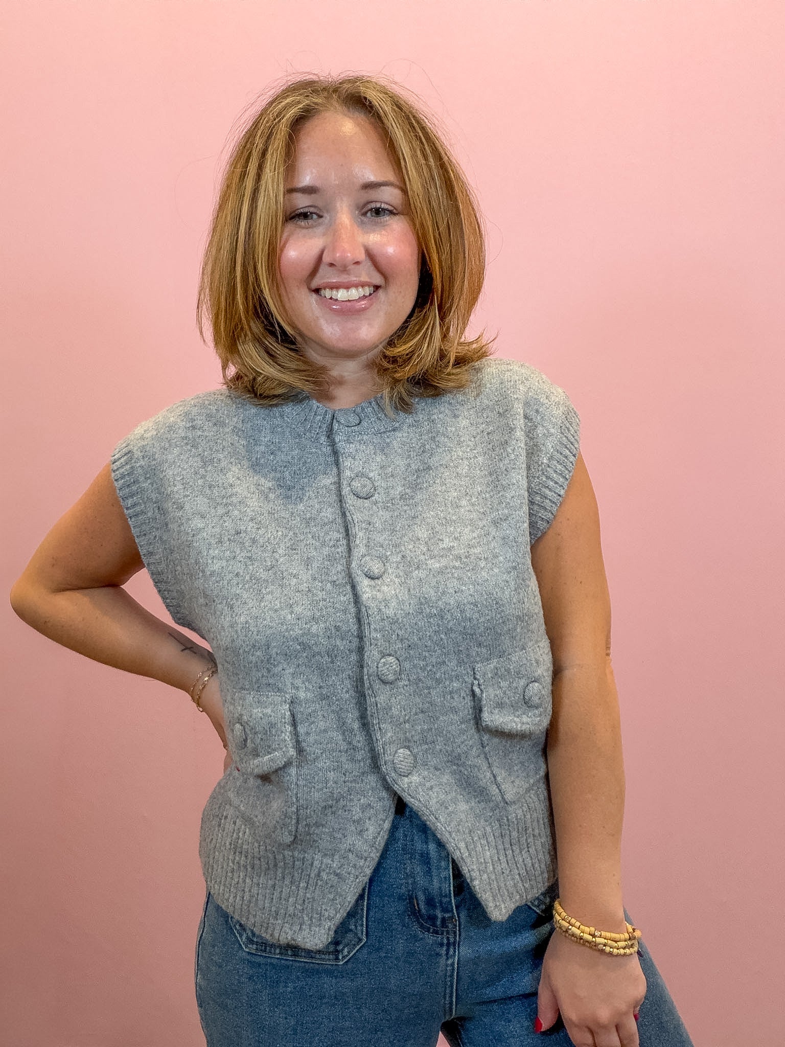 A smiling woman with shoulder-length blonde hair wearing a light gray sleeveless button-front knit vest over a white top and blue jeans, standing against a solid pink background.