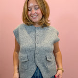 A woman wearing a light gray sleeveless button-front knit vest with front pockets, paired with blue jeans, standing against a solid pink background.