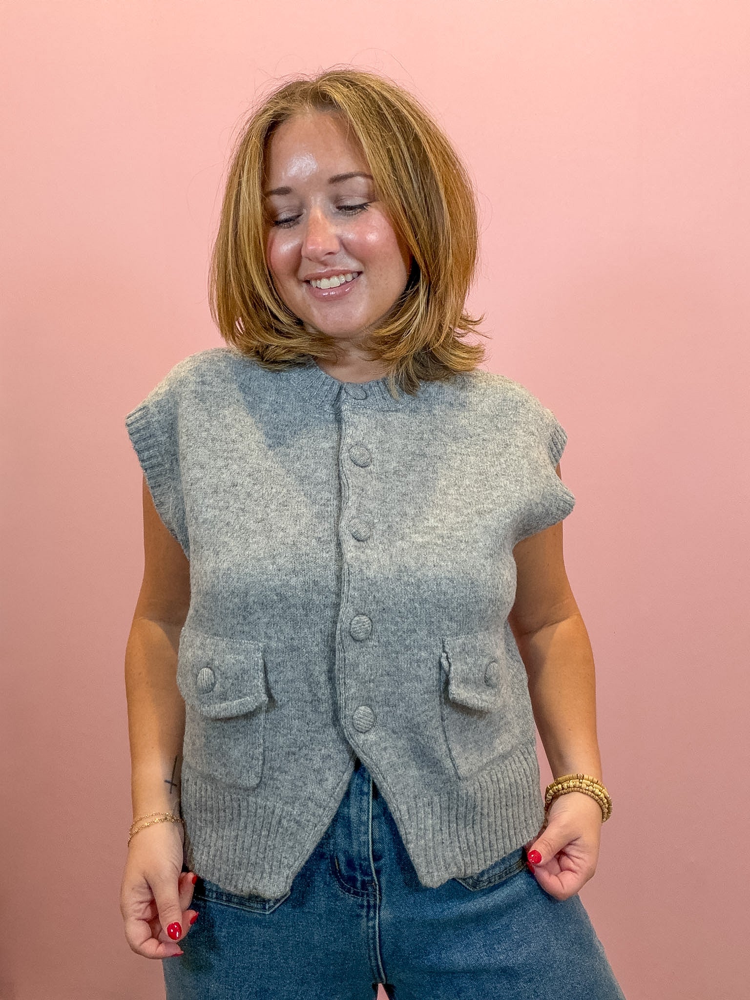 A woman wearing a light gray sleeveless button-front knit vest with front pockets, paired with blue jeans, standing against a solid pink background.