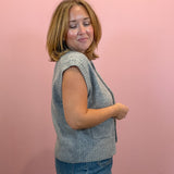 A woman with shoulder-length blonde hair is shown from the side, wearing a light gray ribbed knit sleeveless vest over a dark top and blue jeans, standing against a solid pink background.