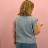 A woman viewed from behind wearing a light gray sleeveless knit top and blue jeans, standing against a solid pink wall.