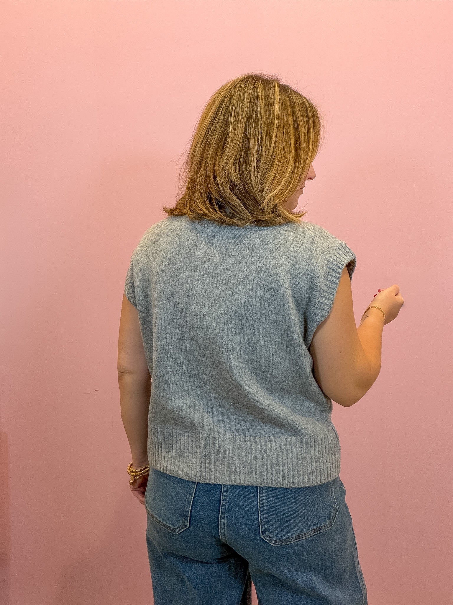 A woman viewed from behind wearing a light gray sleeveless knit top and blue jeans, standing against a solid pink wall.