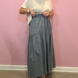 A woman modeling a long tiered skirt with a small white geometric pattern on a slate blue-gray background, paired with a cream t-shirt and woven slide sandals, standing against a pink wall.