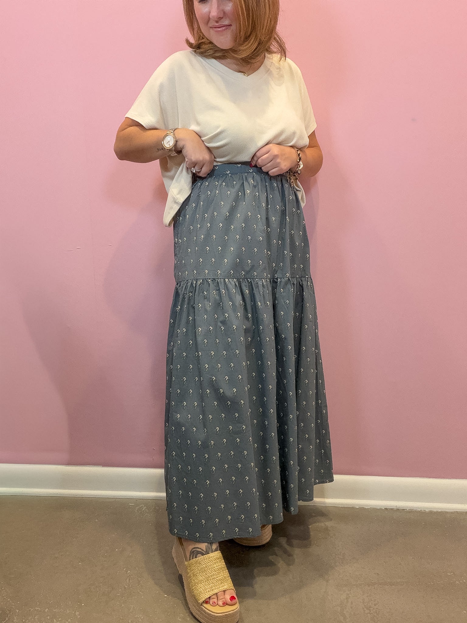 A woman modeling a long tiered skirt with a small white geometric pattern on a slate blue-gray background, paired with a cream t-shirt and woven slide sandals, standing against a pink wall.