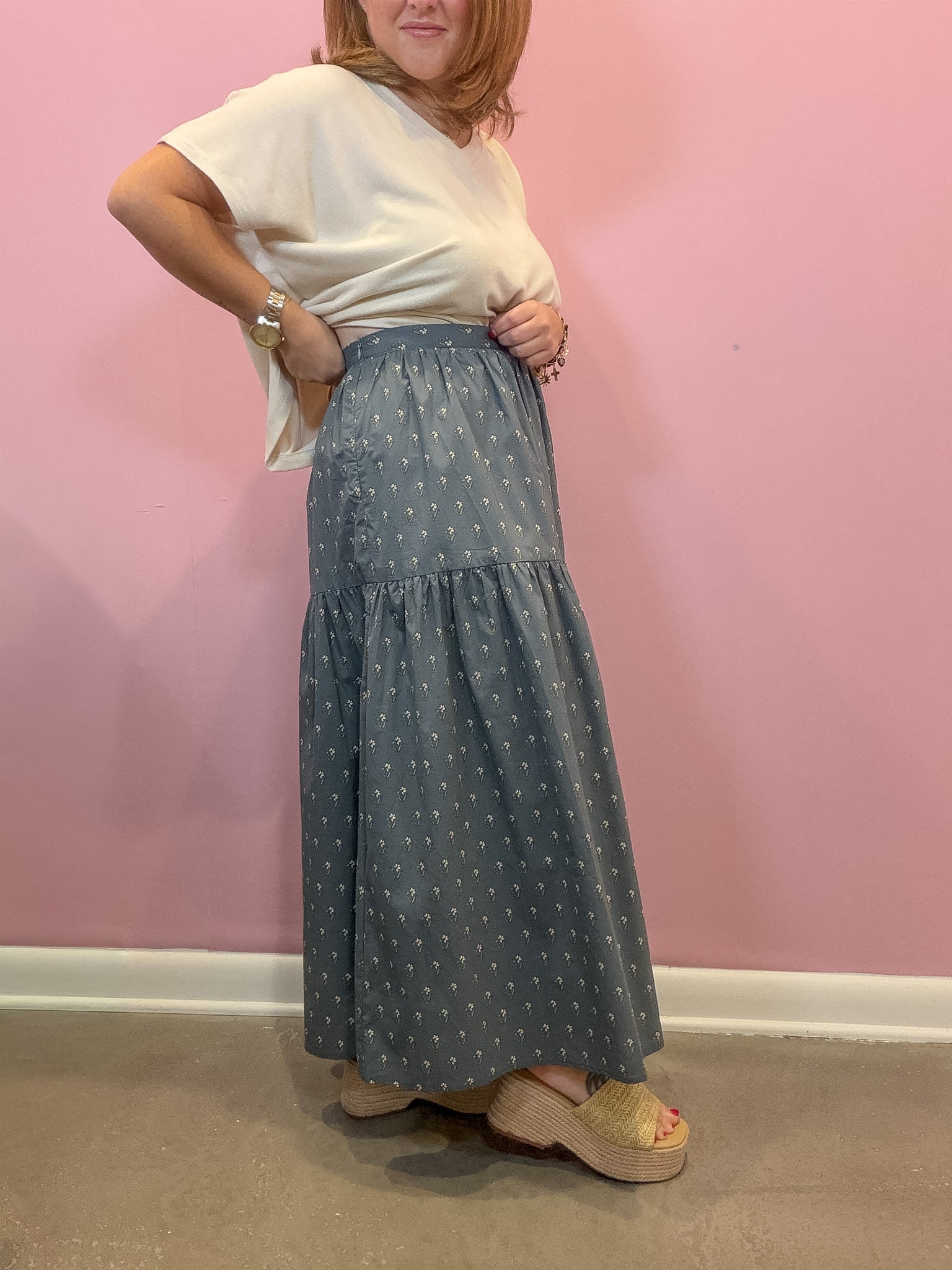 A woman modeling a long tiered skirt in a dusty blue-gray color with a small white polka dot pattern, paired with a cream t-shirt and beige platform sandals, standing against a pink wall.