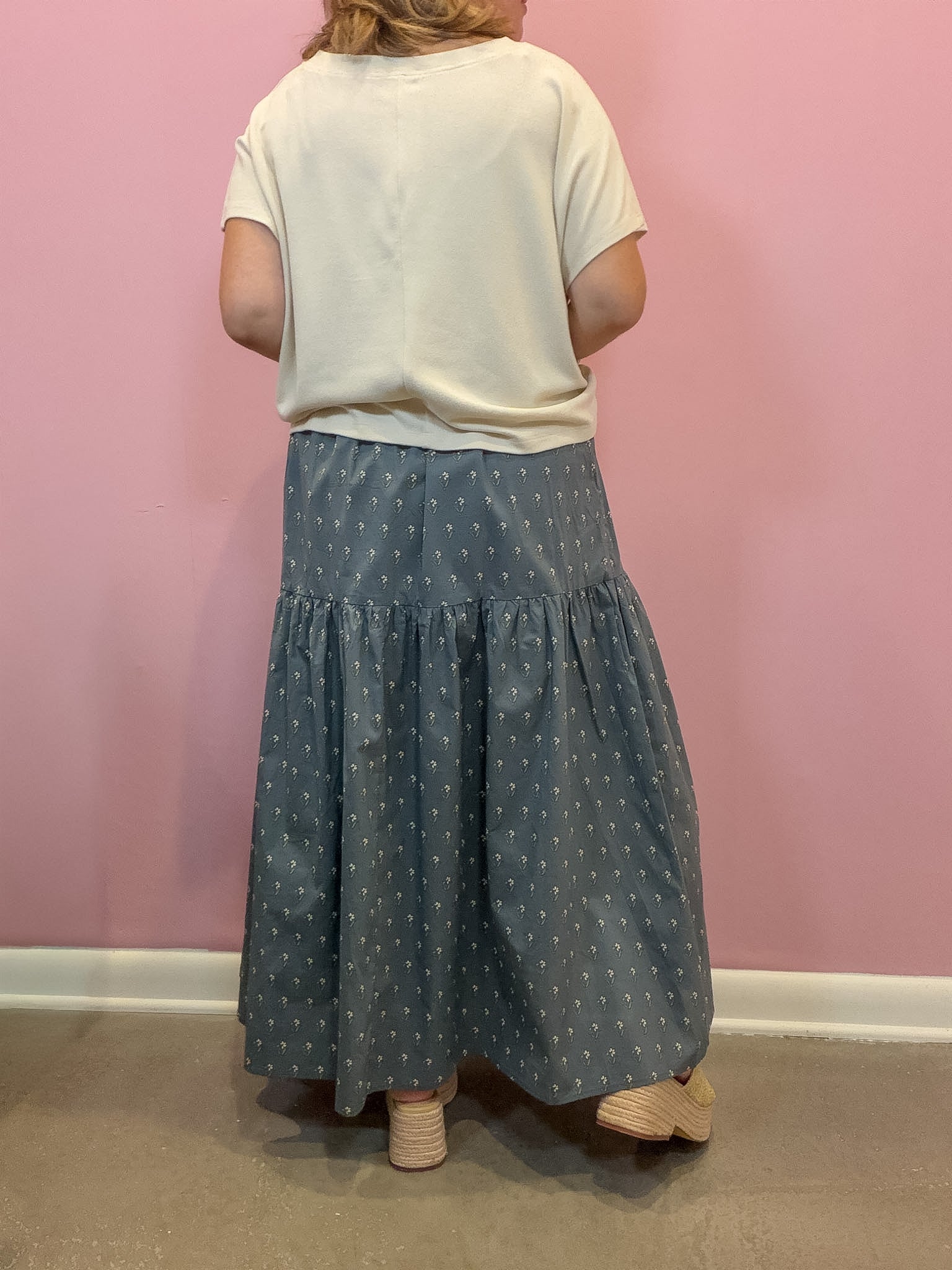 A person viewed from the back wearing a cream short-sleeve top and a tiered slate blue skirt with a small white geometric pattern, standing against a pink wall.