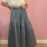 A person wearing a loose white short-sleeve top and a tiered blue-gray maxi skirt with a small white floral print, standing against a pink wall.