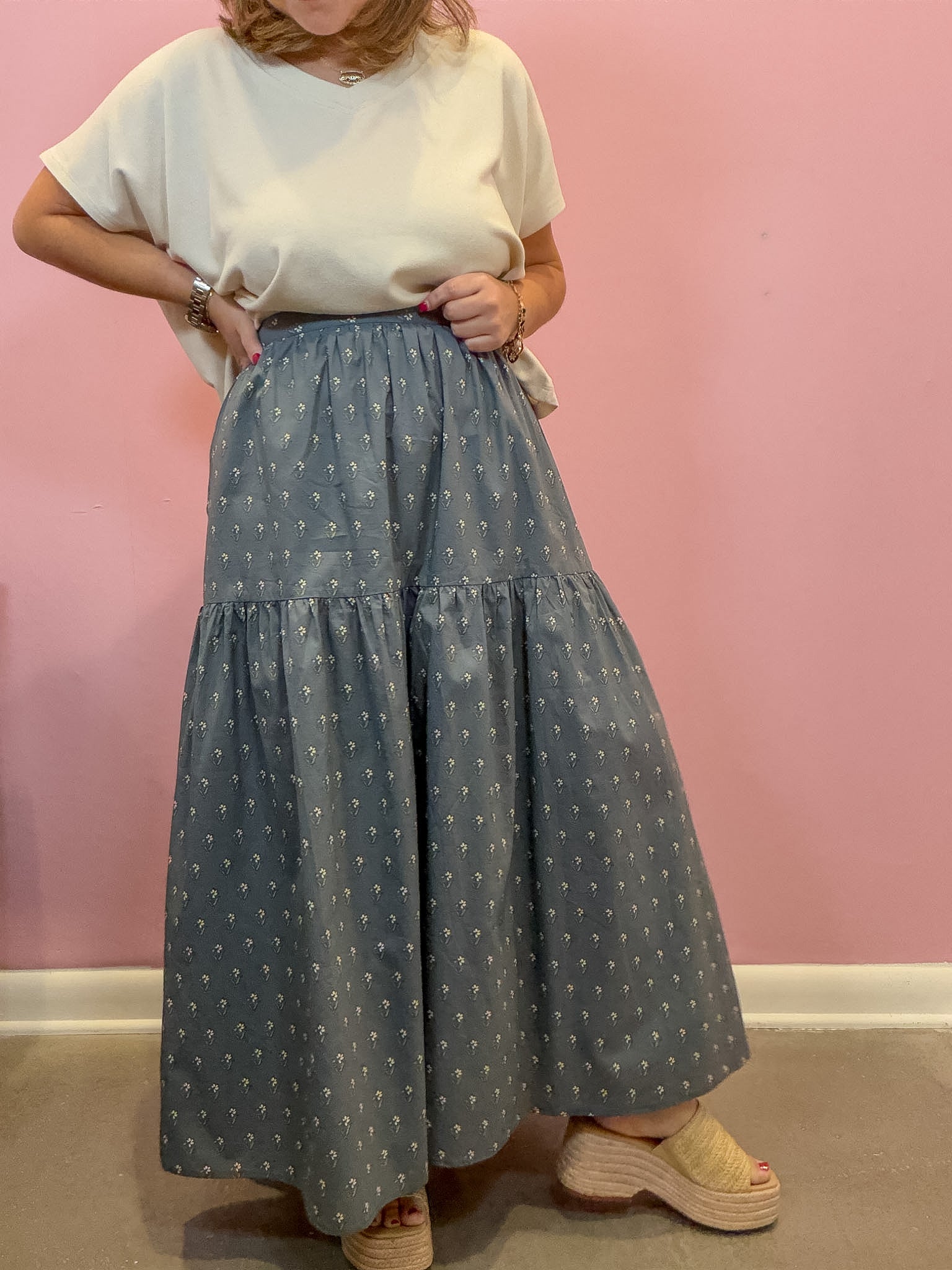 A person wearing a loose white short-sleeve top and a tiered blue-gray maxi skirt with a small white floral print, standing against a pink wall.